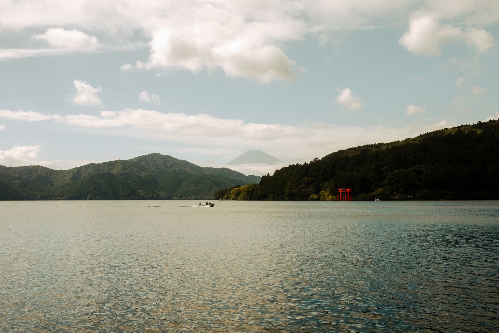 Lake Ashi at Hakone, a small red torii on the right shore, a boat on the water, and Mount Fuji faint in haze between the hills.