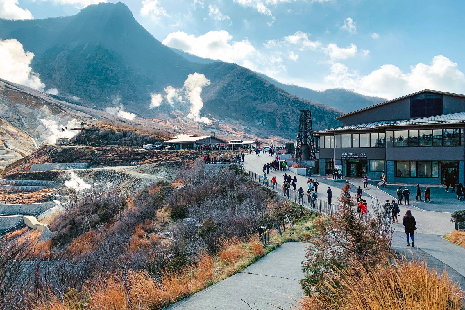 Steam vents on the hillside at Owakudani, Hakone, visitors on the path and a museum building below.