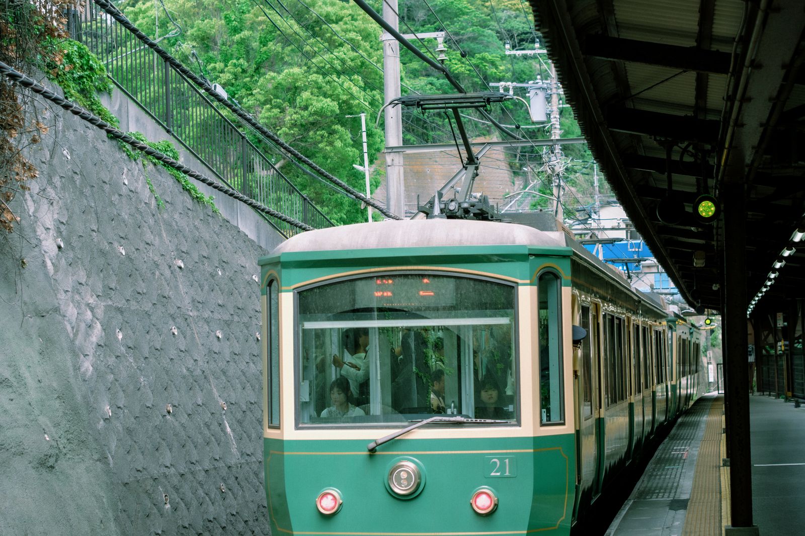 A green Enoden car pulling into a walled station in Kamakura, trees above the wall.