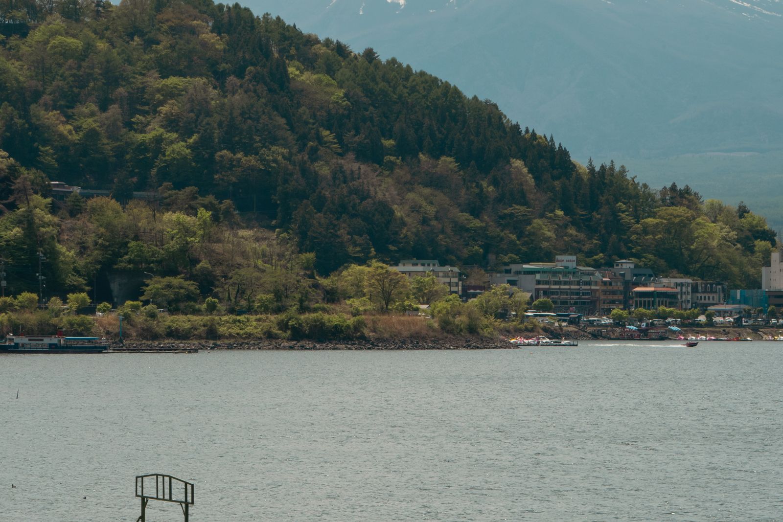 The shore of Lake Kawaguchi with swan boats and the lakeside town, a mountain's lower slope filling the top right of the frame.