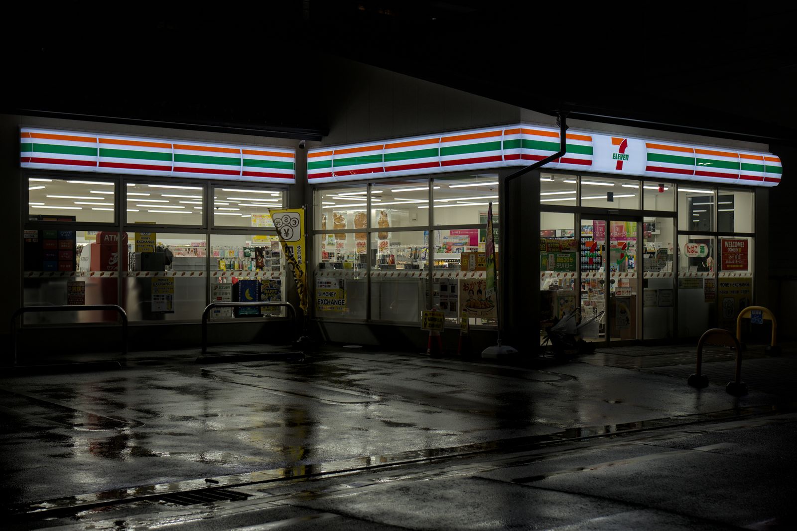 A 7-Eleven in Kyoto lit at night, its fascia reflected in a wet forecourt, money exchange and ATM signs in the window.