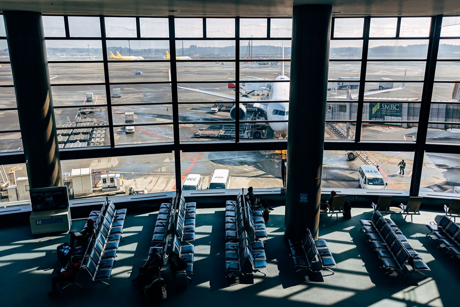 A gate lounge at a Tokyo airport, rows of blue seats in long window shadows, a widebody aircraft at the jet bridge.