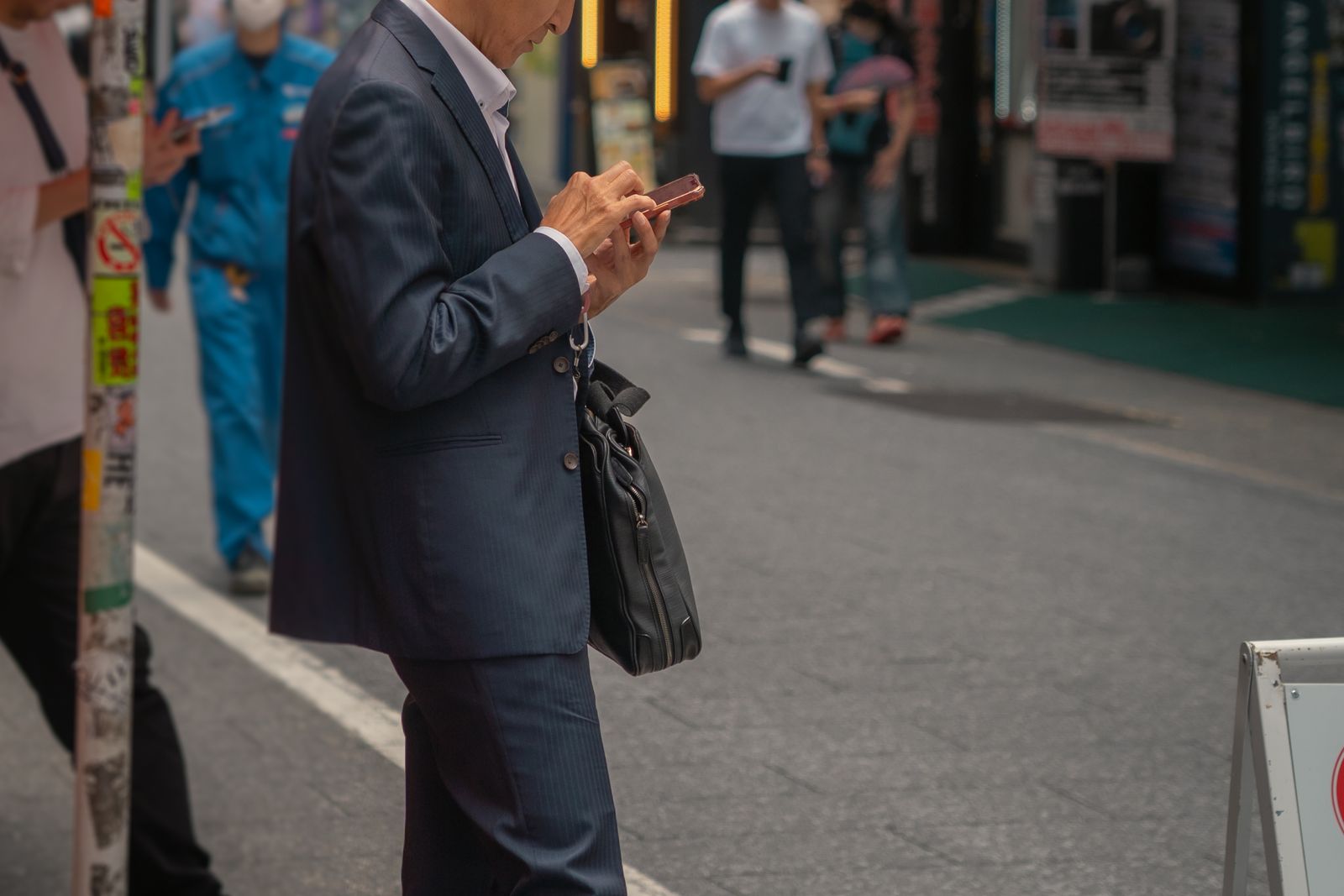 A suited man, face cropped, thumbing a phone at the edge of a Tokyo street, pedestrians blurred behind him.