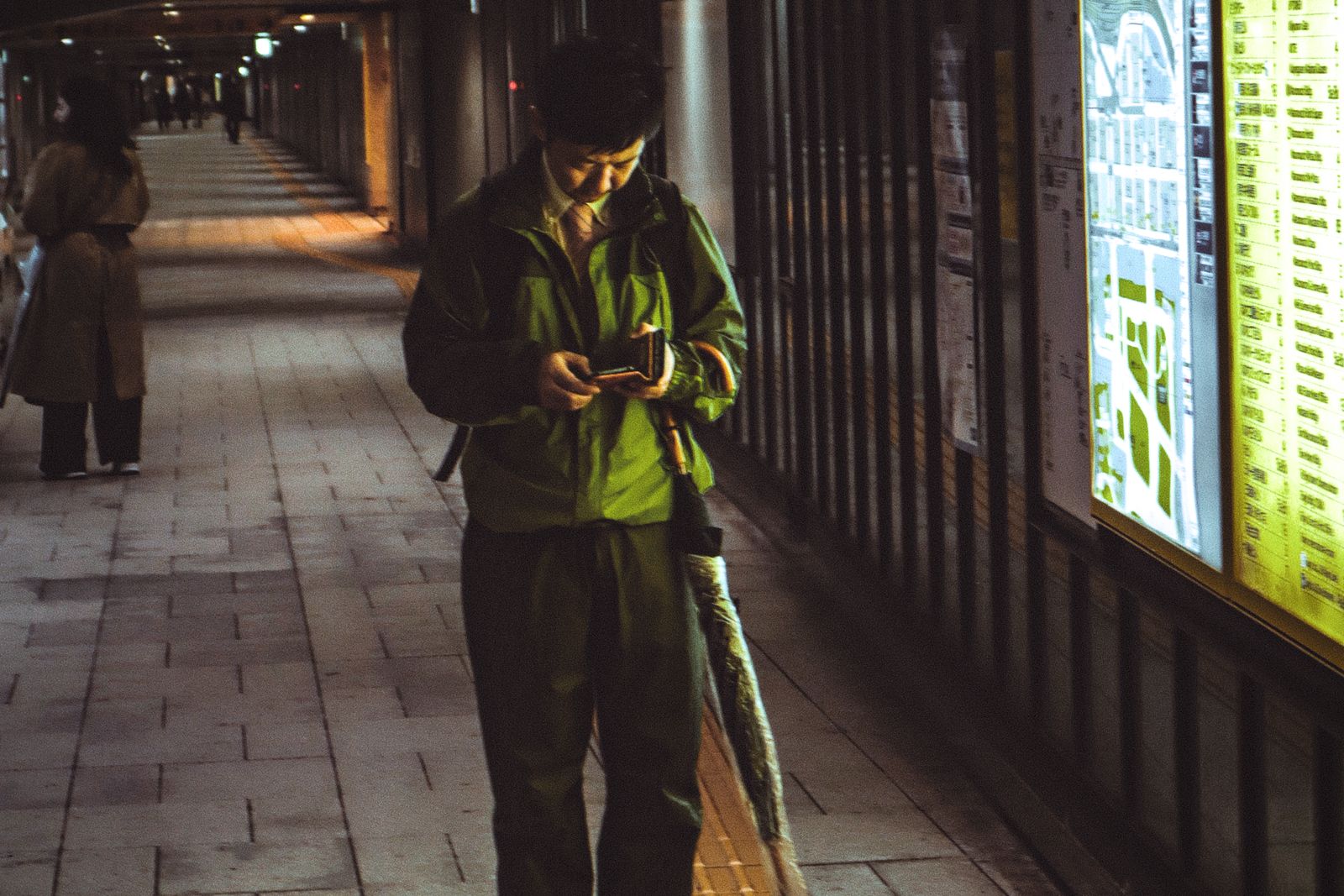 A man with an umbrella walking an underground passage in Tokyo at night, head down over his phone, beside a backlit area-map board.