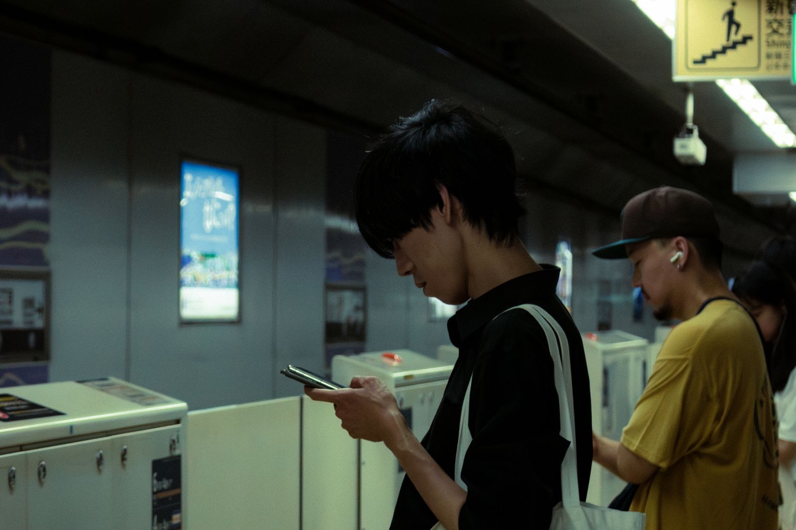 A young man in profile reading his phone beside the ticket gates of a Tokyo subway station, a stairs pictogram sign in Japanese overhead.