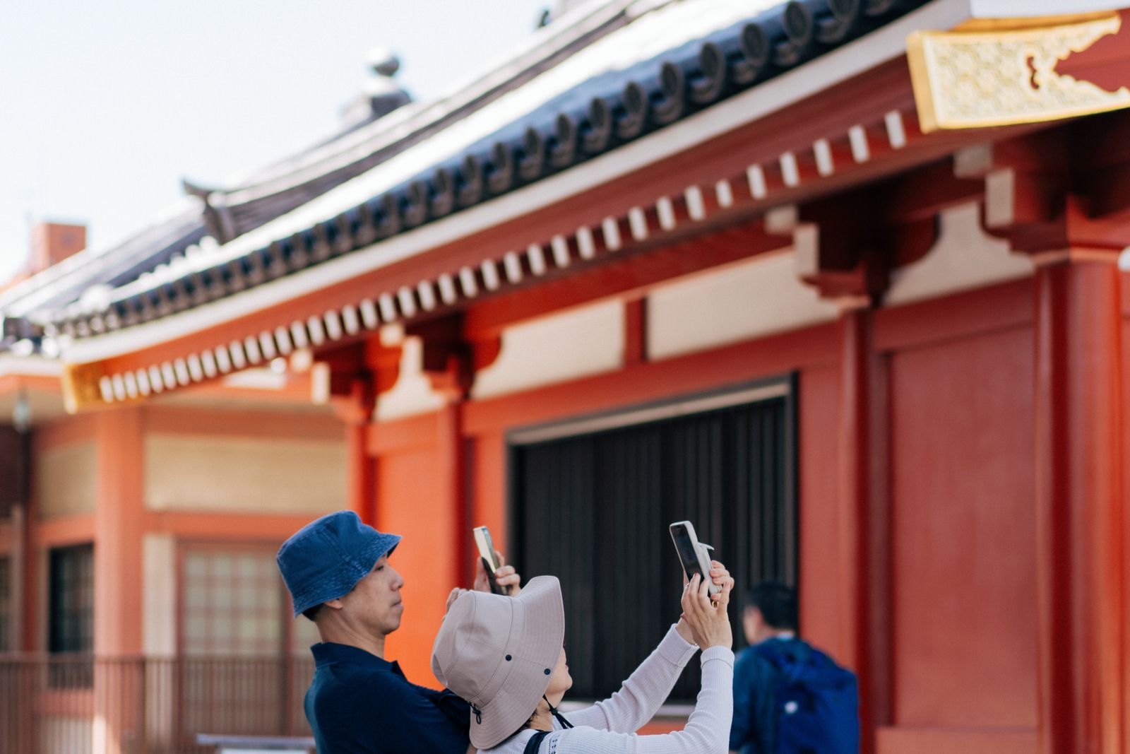 Three visitors in front of a vermilion temple hall in Tokyo, two of them holding phones up to photograph it.