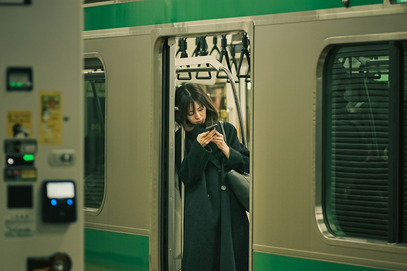 A woman in a dark coat in the open doorway of a green-striped commuter train at Shinagawa, head down over her phone.