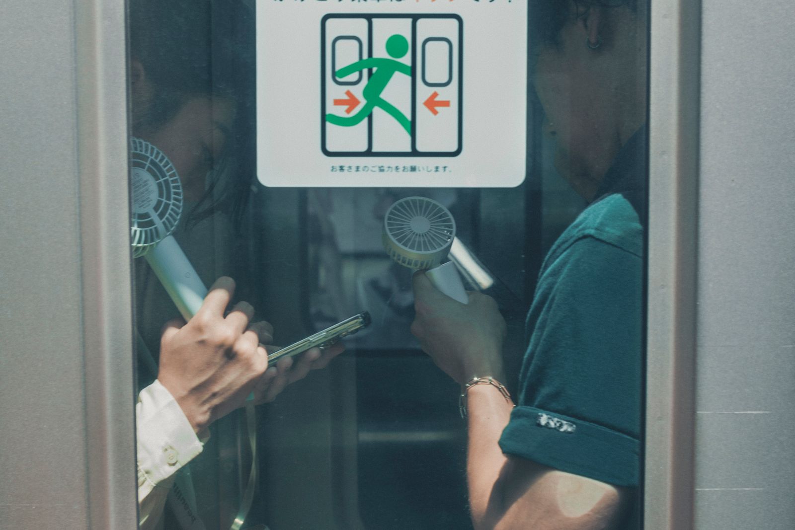 Two passengers seen through a train door window in Toshima, Tokyo, each holding a handheld fan, one thumbing a phone.
