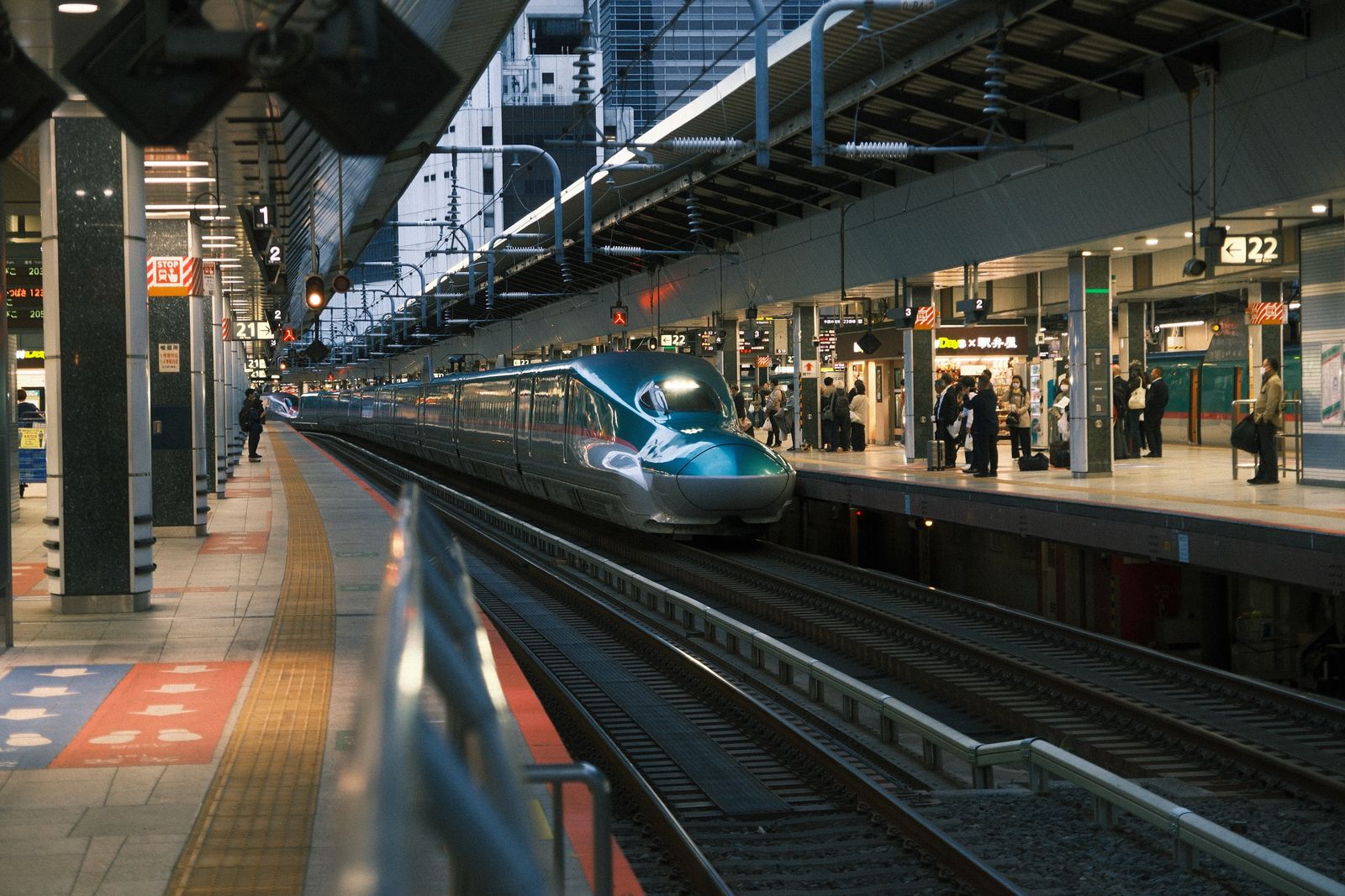 A shinkansen platform at Tokyo Station in the evening, with the dark green front car of a train stopped alongside it. On the platform opposite, passengers wait to depart and a station bento shop is lit.