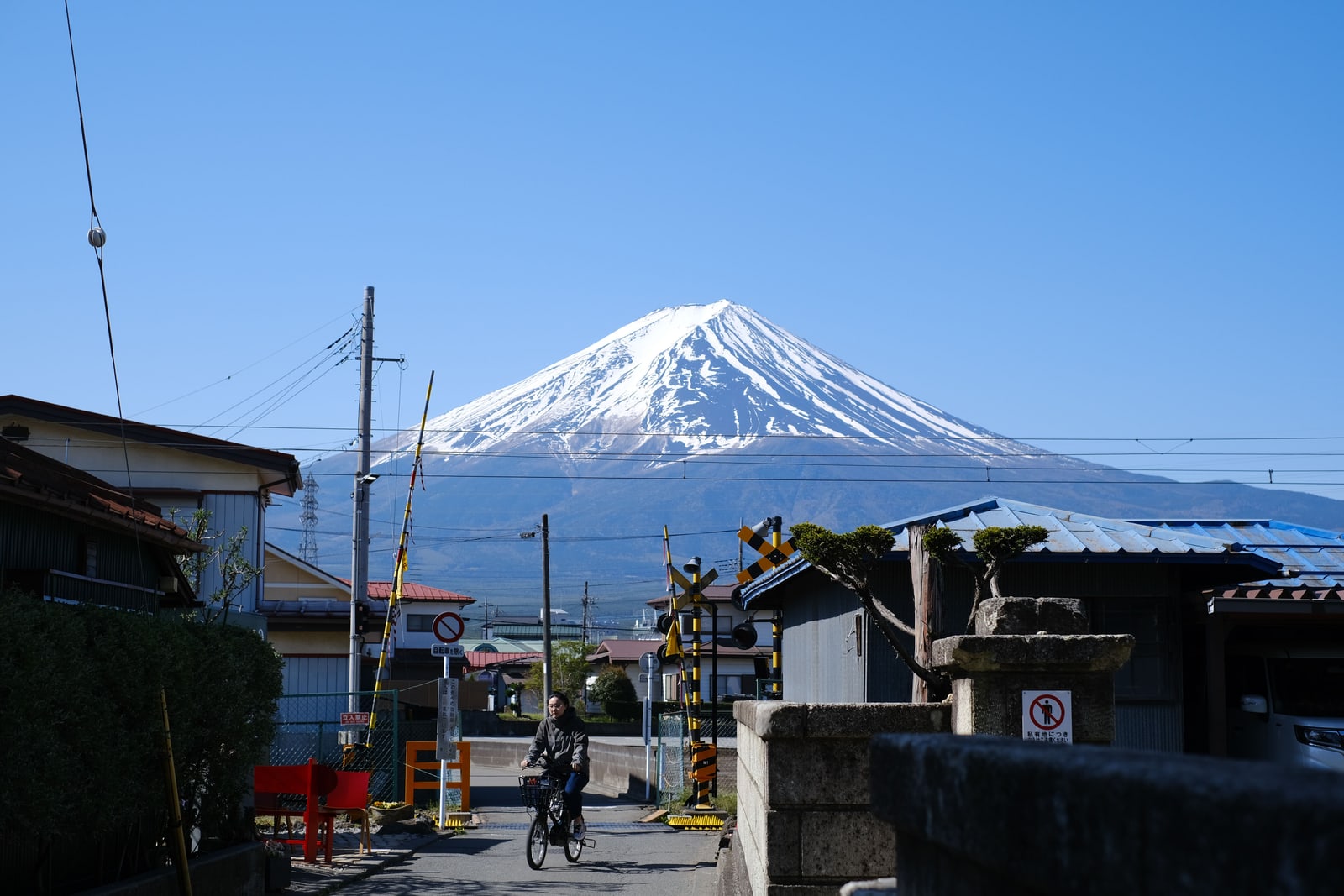 A snow-capped Mount Fuji under a clear blue sky behind a small-town railway crossing, with a cyclist on the road.