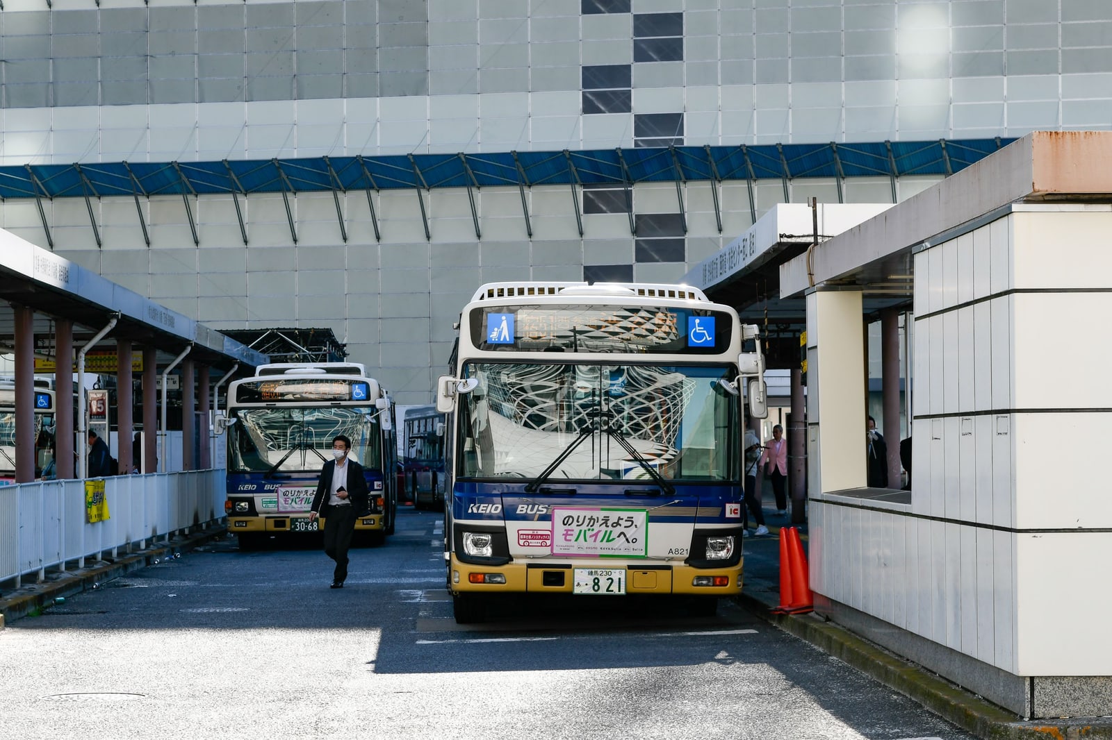 Two Keio buses at the bays of an open-air bus terminal, with a passenger walking past.