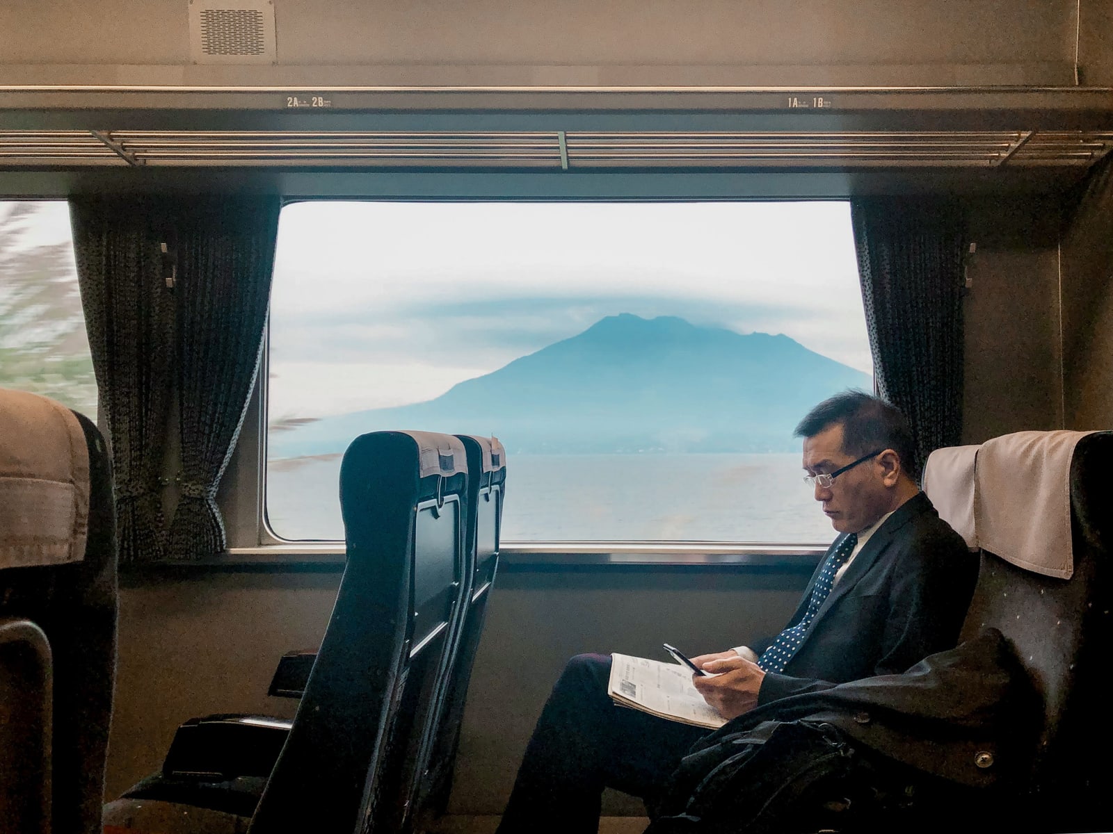 A man in a dark suit reading in a reclining train seat beside a wide window, with a mountain across water in haze outside.