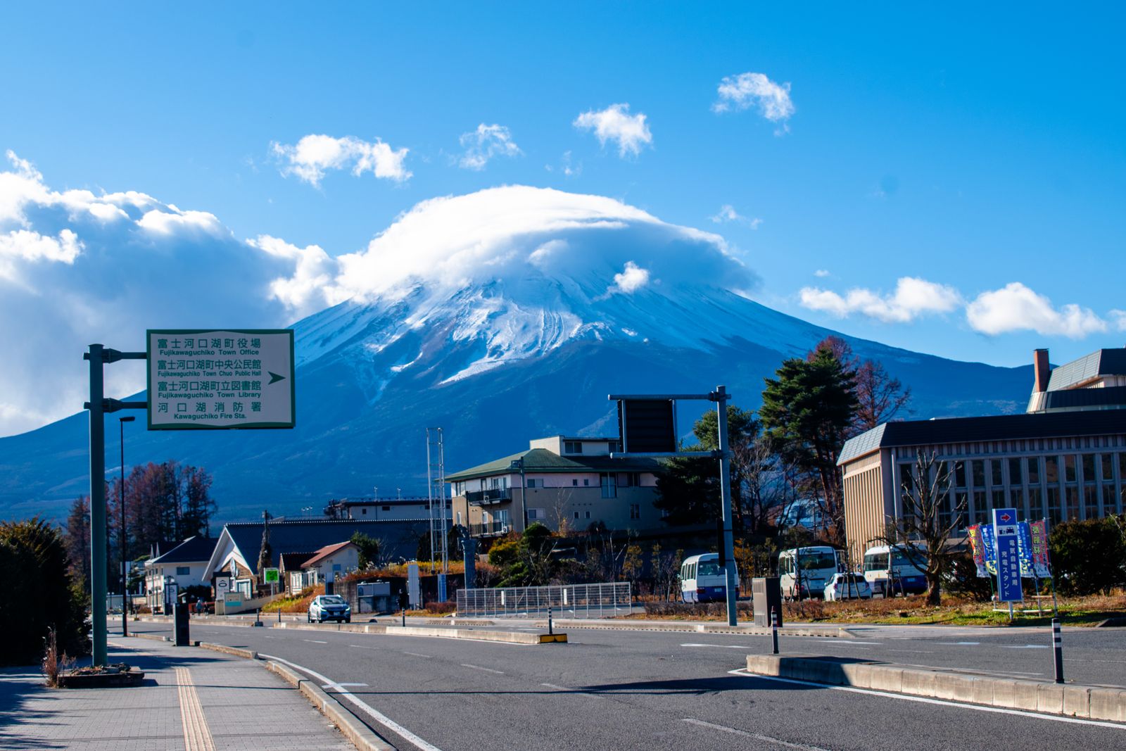 Mount Fuji with a lens-shaped cloud on its summit above a town road in Fujikawaguchiko, with a green direction sign.