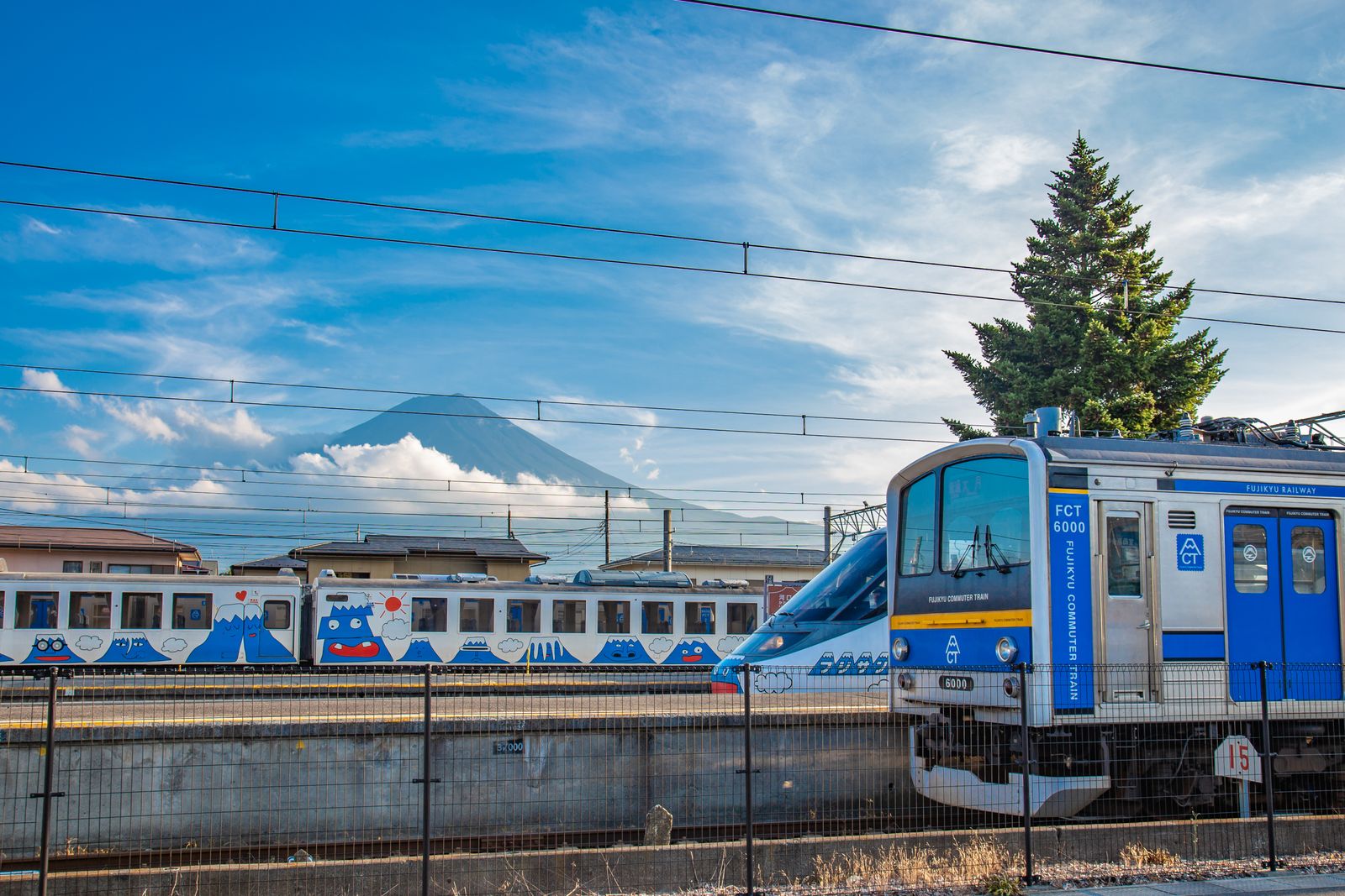 A blue-and-silver Fujikyu Railway train and the nose of a Fujisan View Express at a station, Mount Fuji half in cloud behind the overhead wires.