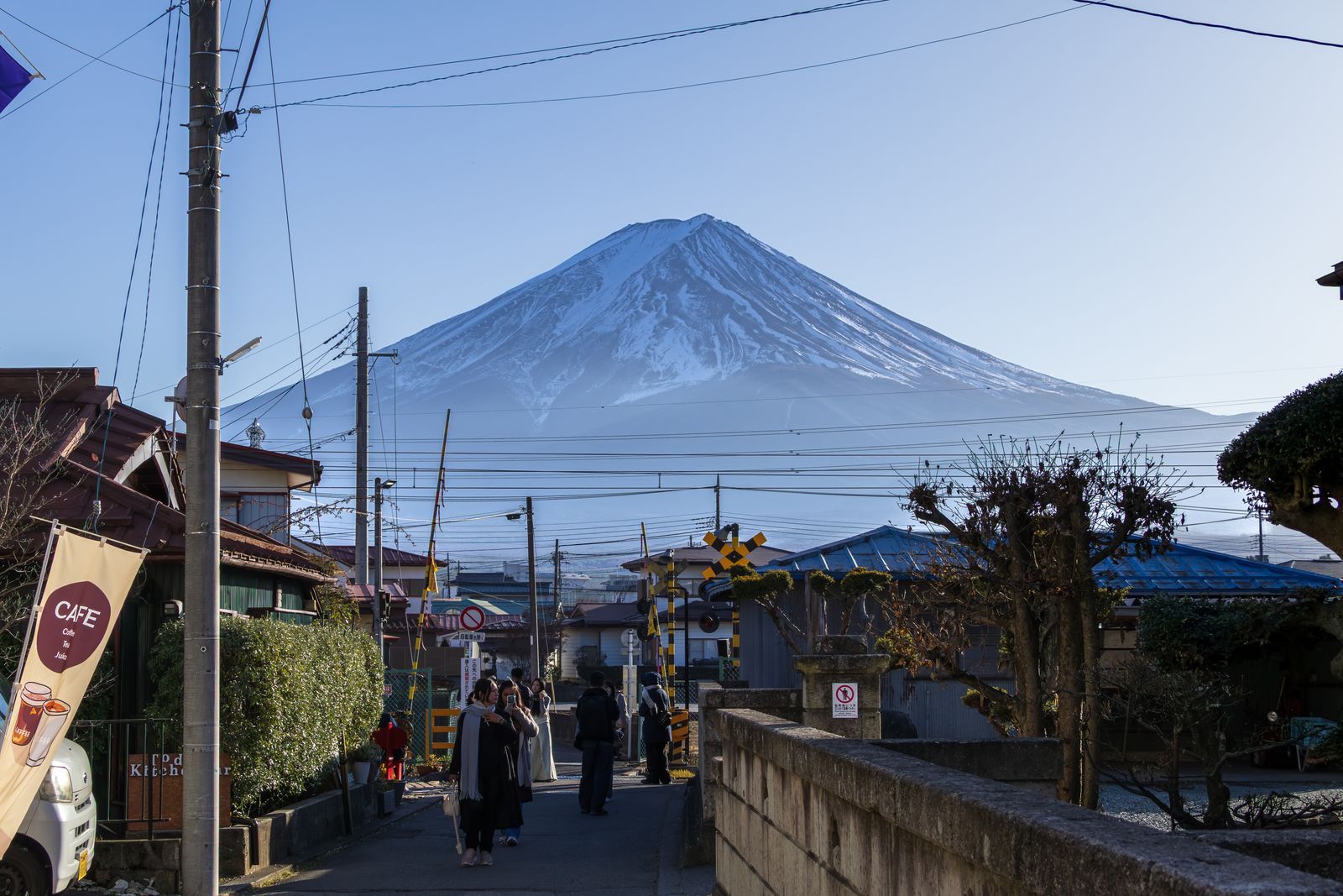 A narrow street in Fujiyoshida leading to a railroad crossing, people walking toward it, Mount Fuji rising above the rooftops and power lines.