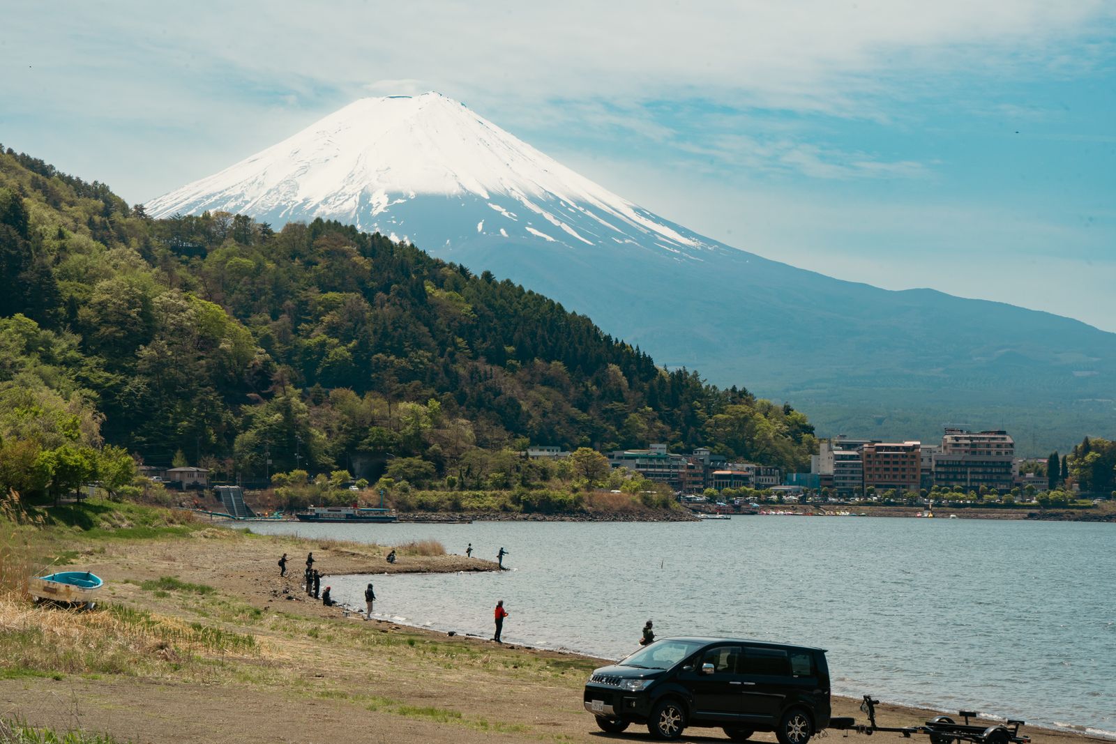Snow-capped Mount Fuji above a wooded ridge and the lakeside hotels of Kawaguchiko, anglers along the near shore of Lake Kawaguchi.