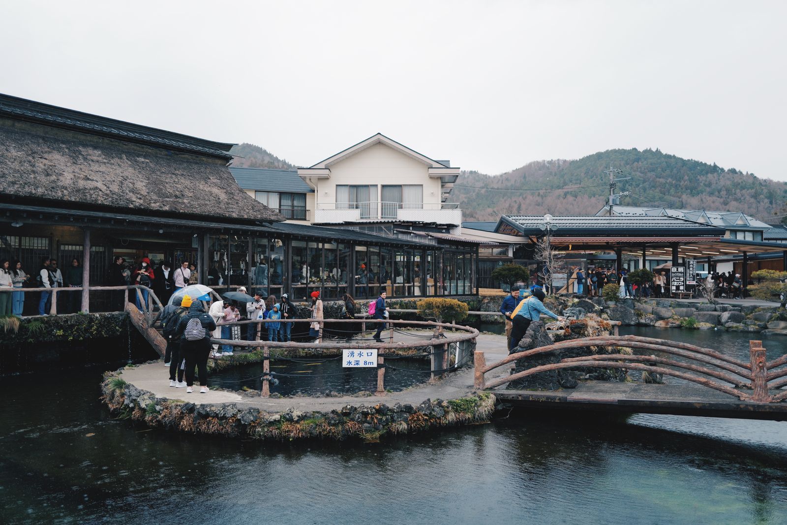 Visitors, some with umbrellas, around a railed spring pond at Oshino Hakkai under a gray sky, a thatched building and souvenir stalls behind, a sign reading spring outlet, depth 8 m.
