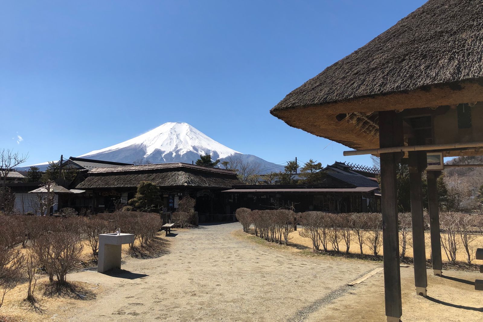 Thatched-roof buildings and a gravel path at Oshino under a clear blue sky, a snow-capped Mount Fuji rising behind them.