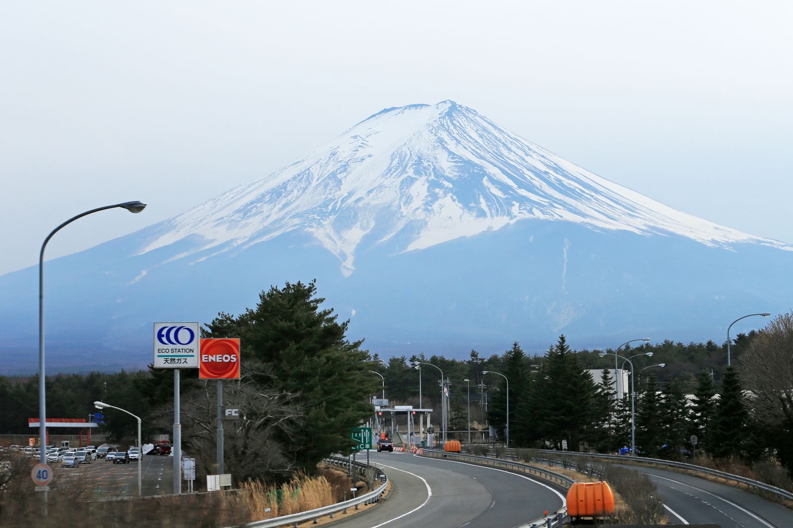 A snow-covered Mount Fuji above an expressway interchange, pine trees and a roadside gas-station sign in the foreground.