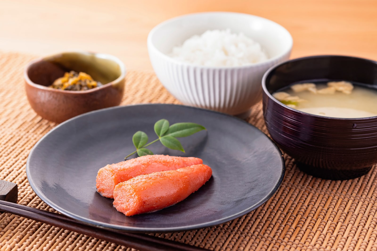 Two lobes of karashi mentaiko on a dark plate beside a bowl of white rice, miso soup and pickles.