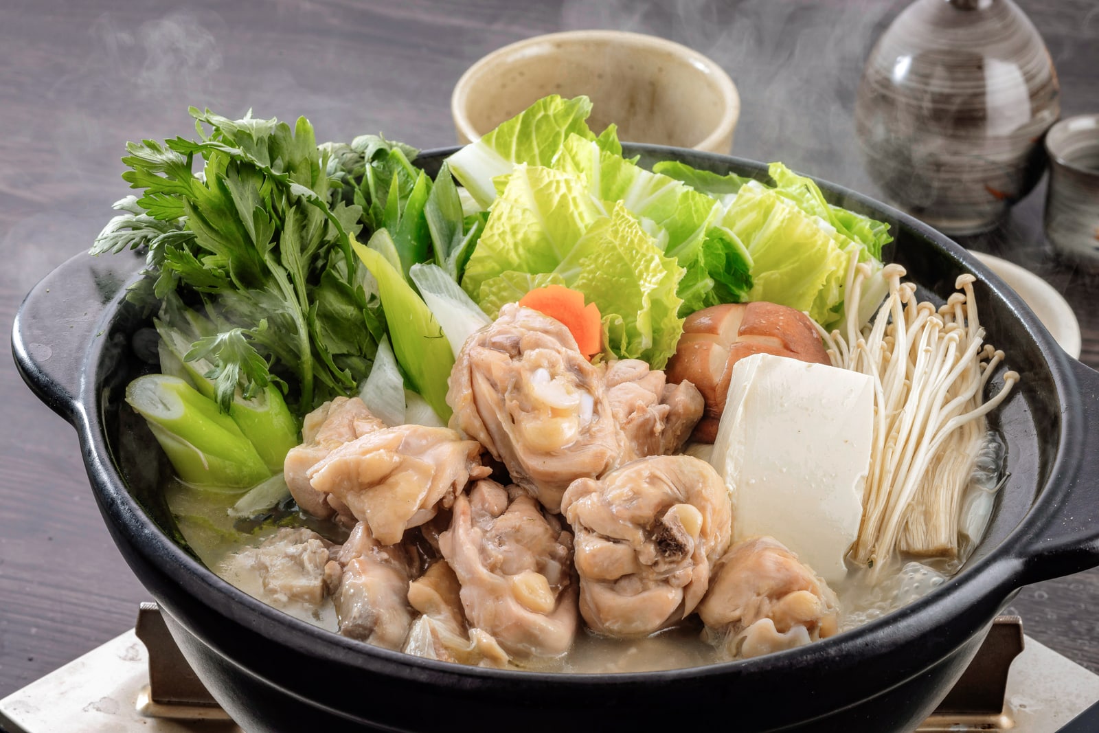 A clay pot of mizutaki — chicken pieces on the bone in a pale broth with tofu, enoki, leeks and greens — set on a table burner.