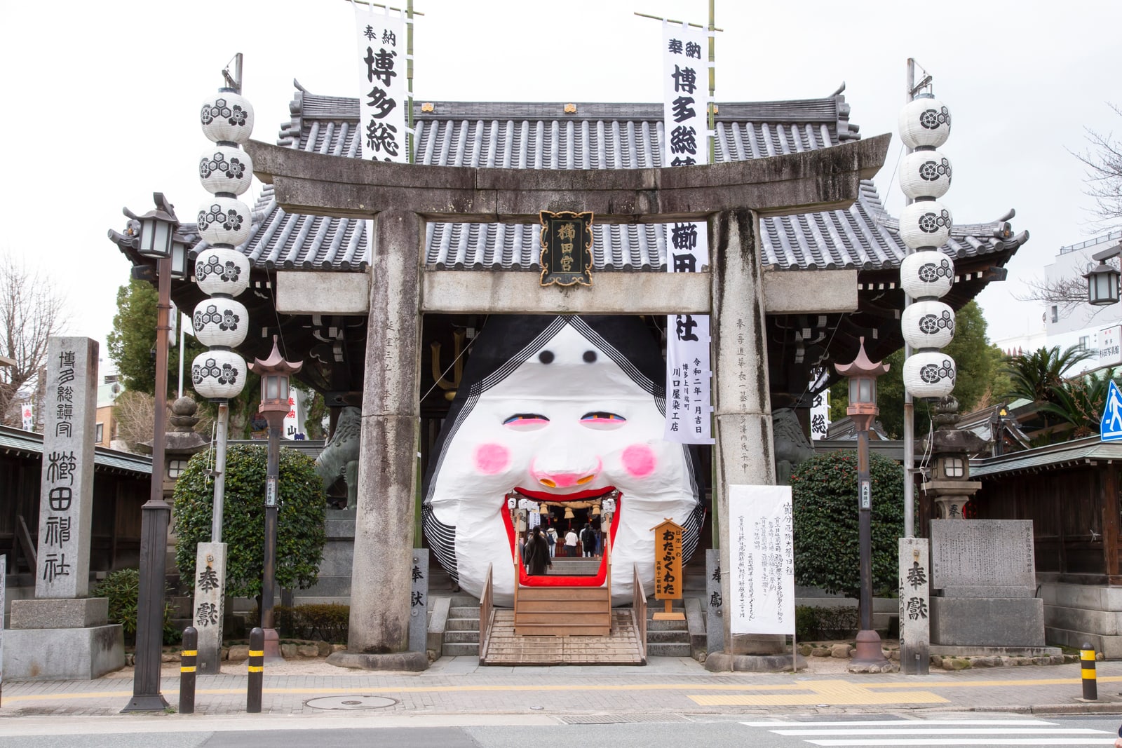 A very large white Otafuku face built over the front of a shrine hall behind a stone torii, with stacked white lanterns and festival banners, set up for Setsubun