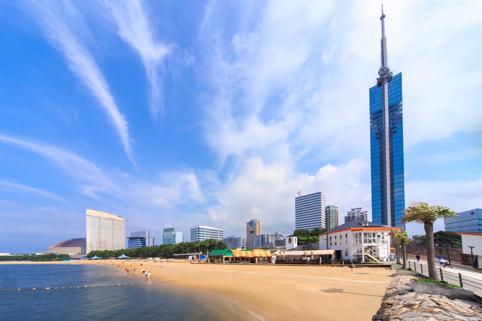 A tall blue-glass tower rising behind a sand beach with a few people at the water edge and small beach buildings, a domed stadium on the left