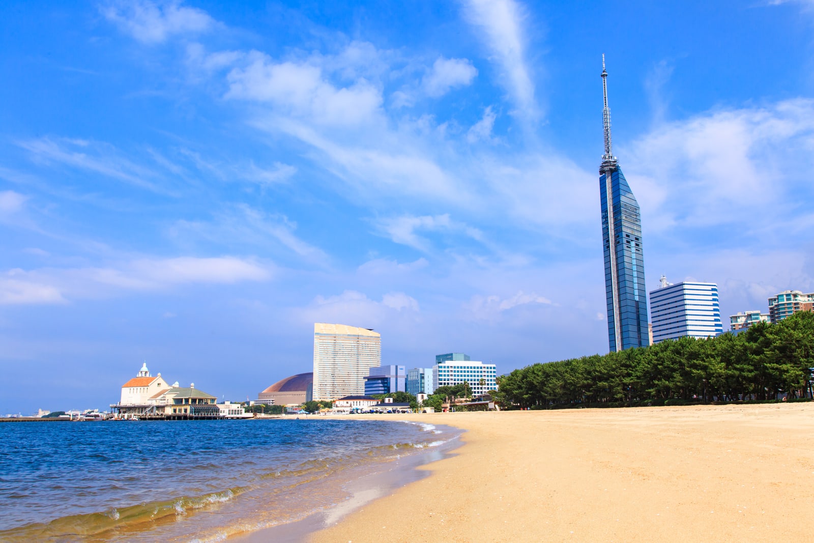 An empty sand beach with gentle waves, a tall blue-glass tower on the right behind a line of pines and a pier building with a green roof on the far left