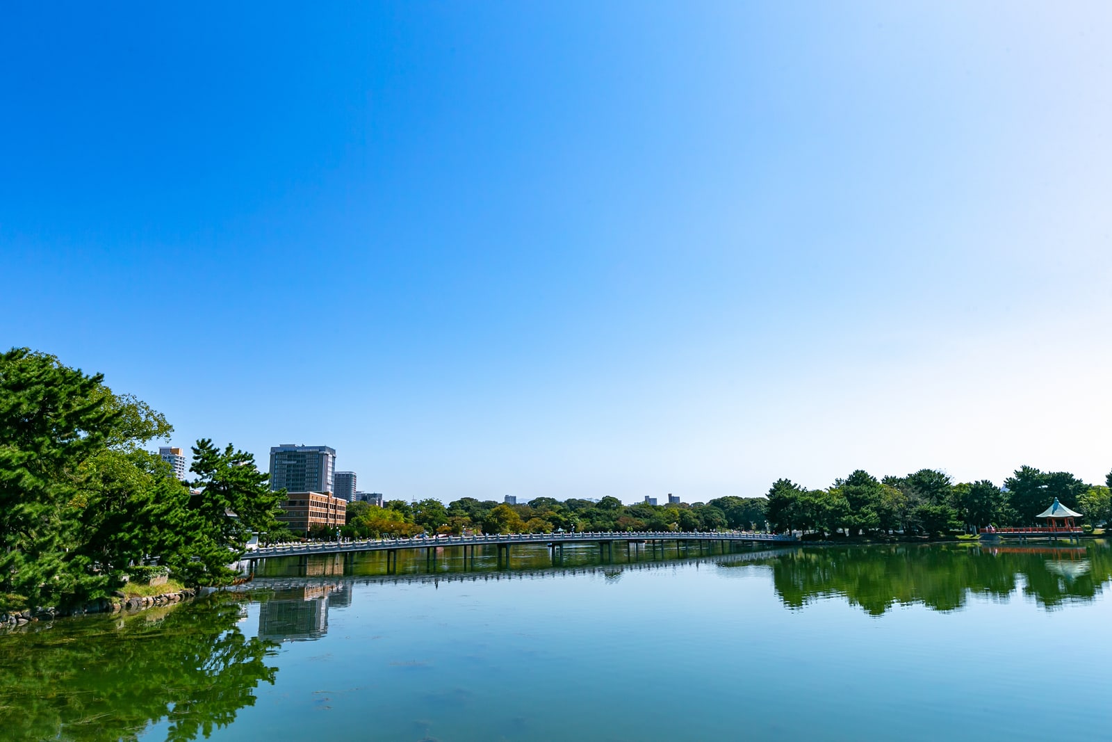 A pond in clear weather with a long low bridge crossing it, a red-roofed pavilion on the right bank and trees reflected in the water