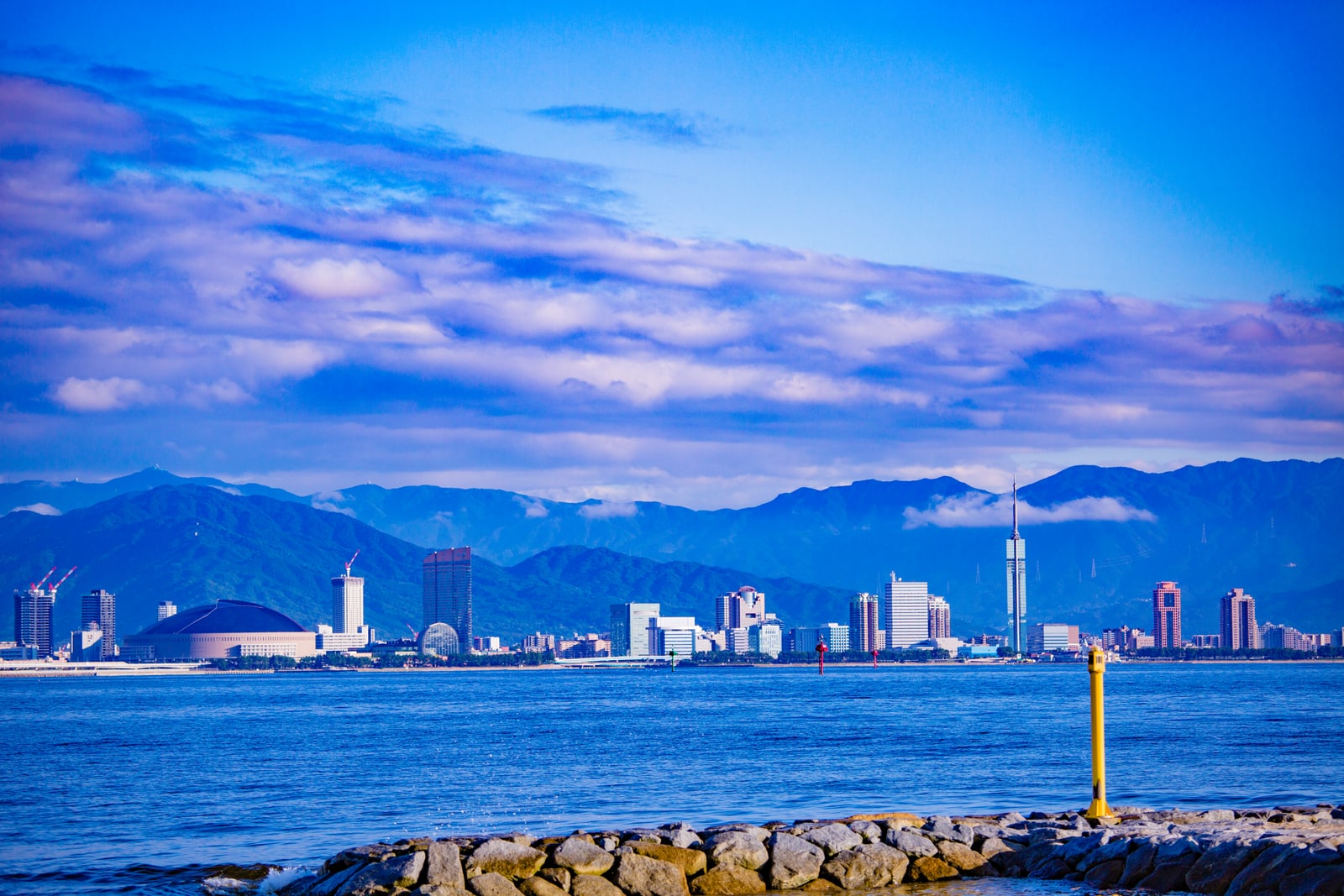 A city skyline across a bay seen from a rock breakwater with a yellow marker post, a domed stadium on the left, a slim tall tower on the right and mountains behind