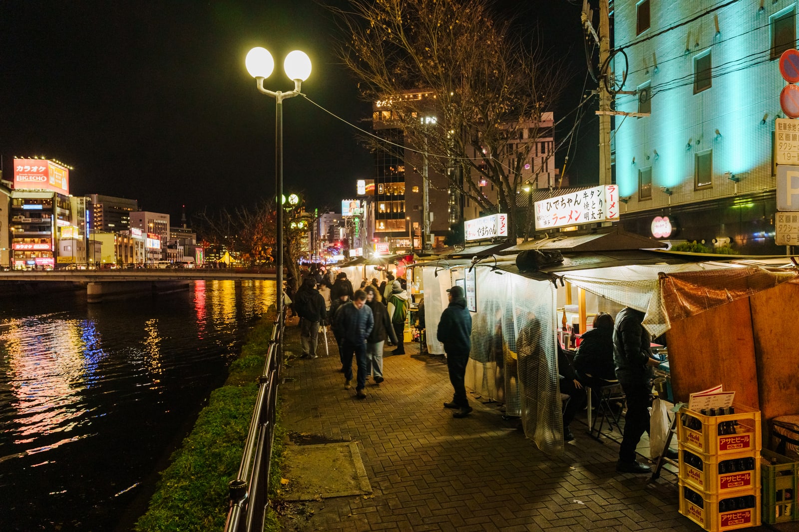 A row of yatai lit along the Nakasu riverbank at night — customers at the counters and queuing on the pavement, the canal and the city's neon beyond.