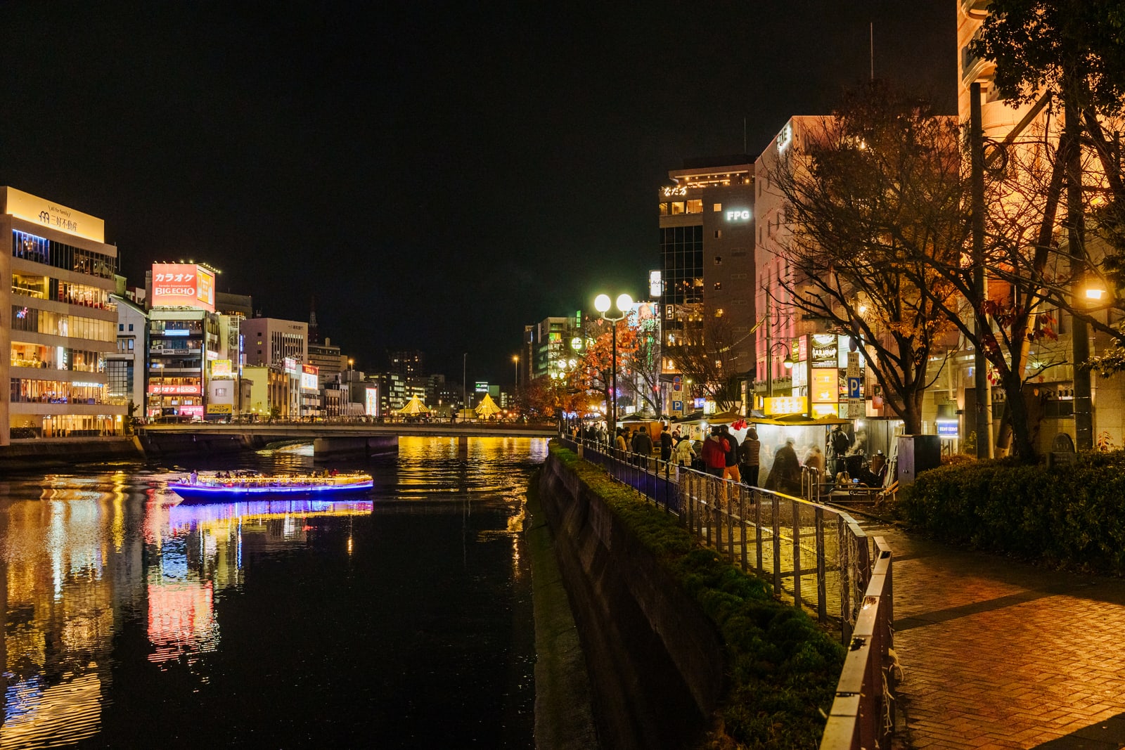 The Nakasu canal at night from the bank, a lit row of yatai along the right-hand promenade and an illuminated boat on the water.