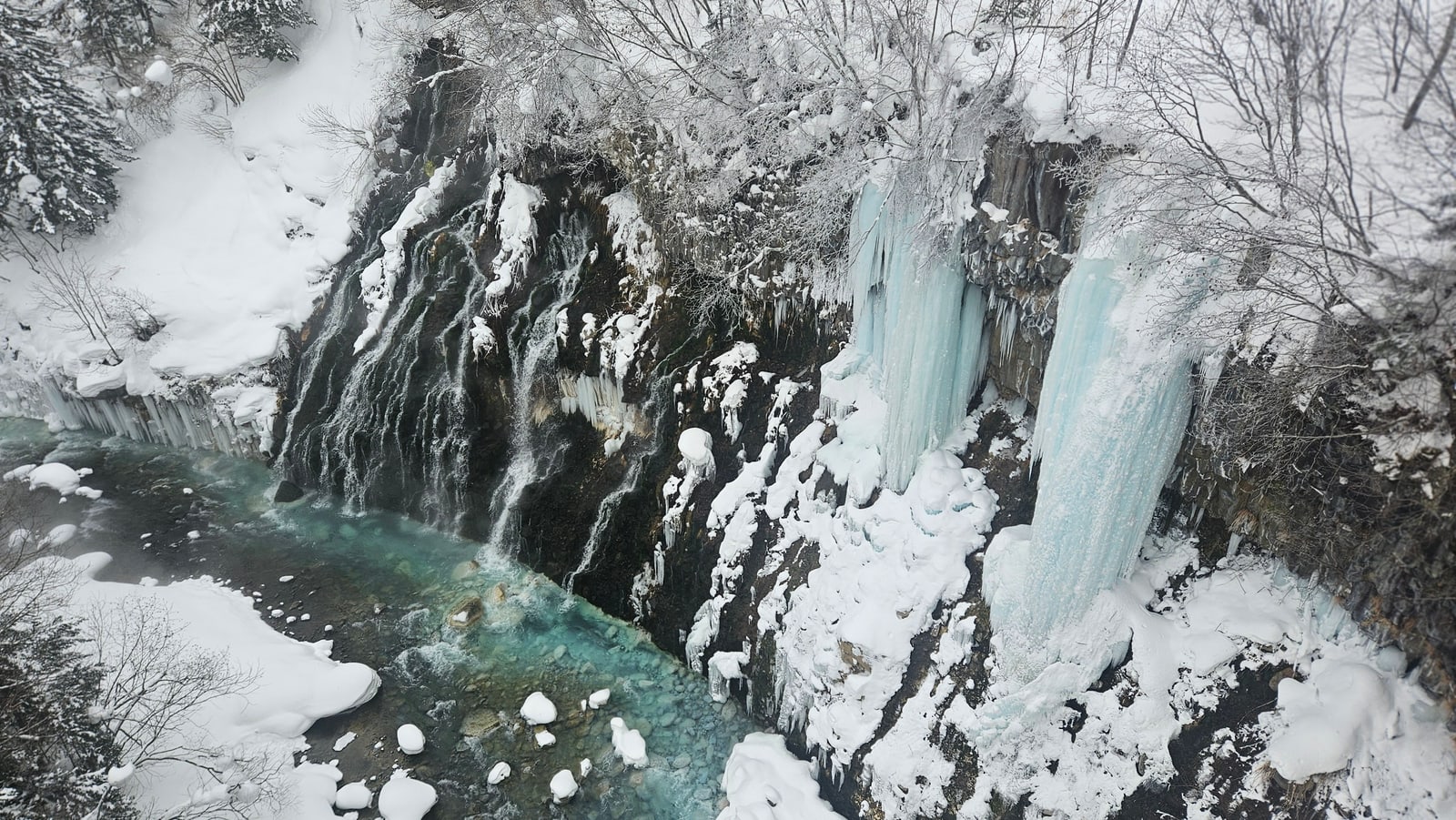 A dark rock face with icicles and snow above a turquoise river, seen from above