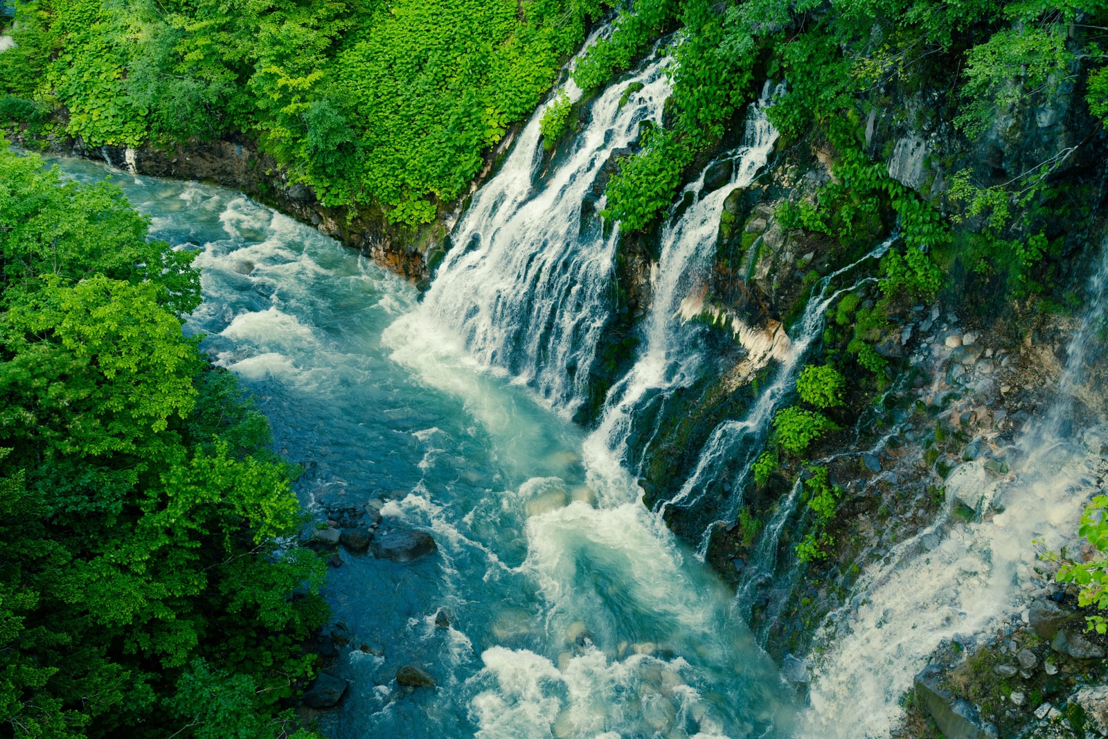 White water falling down a rock face lined with green leaves into a turquoise river, seen from above