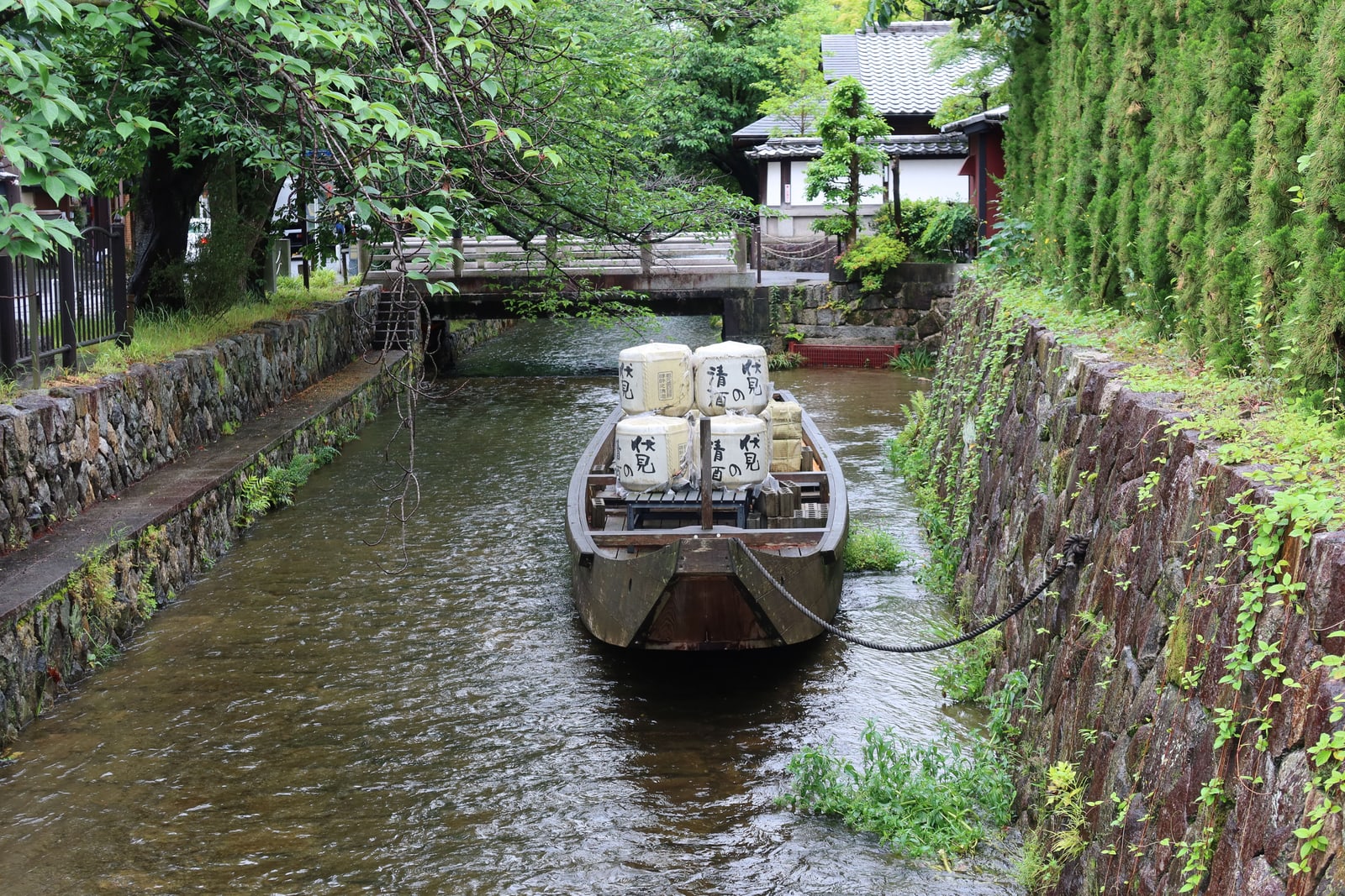 A wooden boat loaded with sake barrels marked 伏見 清酒 in a narrow stone-walled canal under green trees