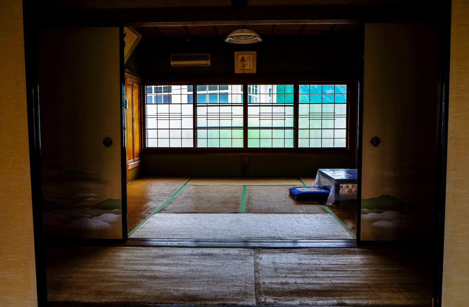 A Japanese room with tatami flooring, shoji windows and painted fusuma doors, empty except for a low table and a floor cushion.
