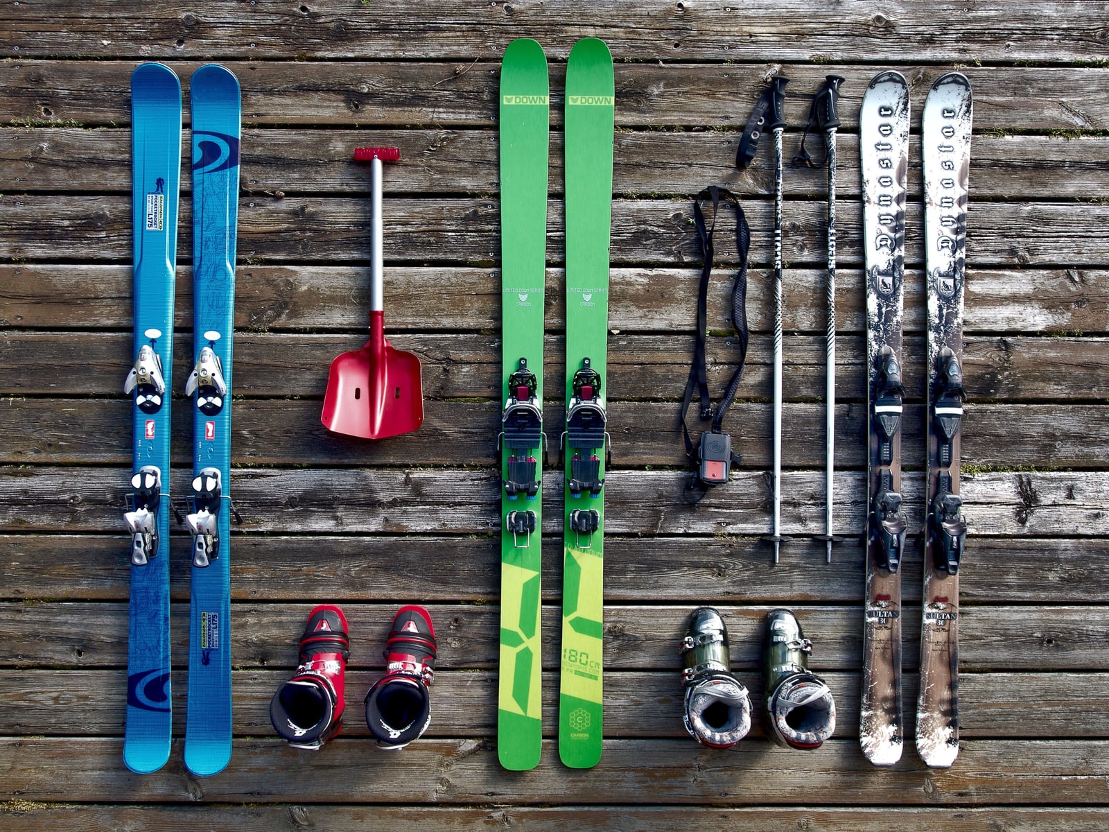 Three pairs of skis, two pairs of ski boots, poles and a red snow shovel laid out on dark wooden planks.