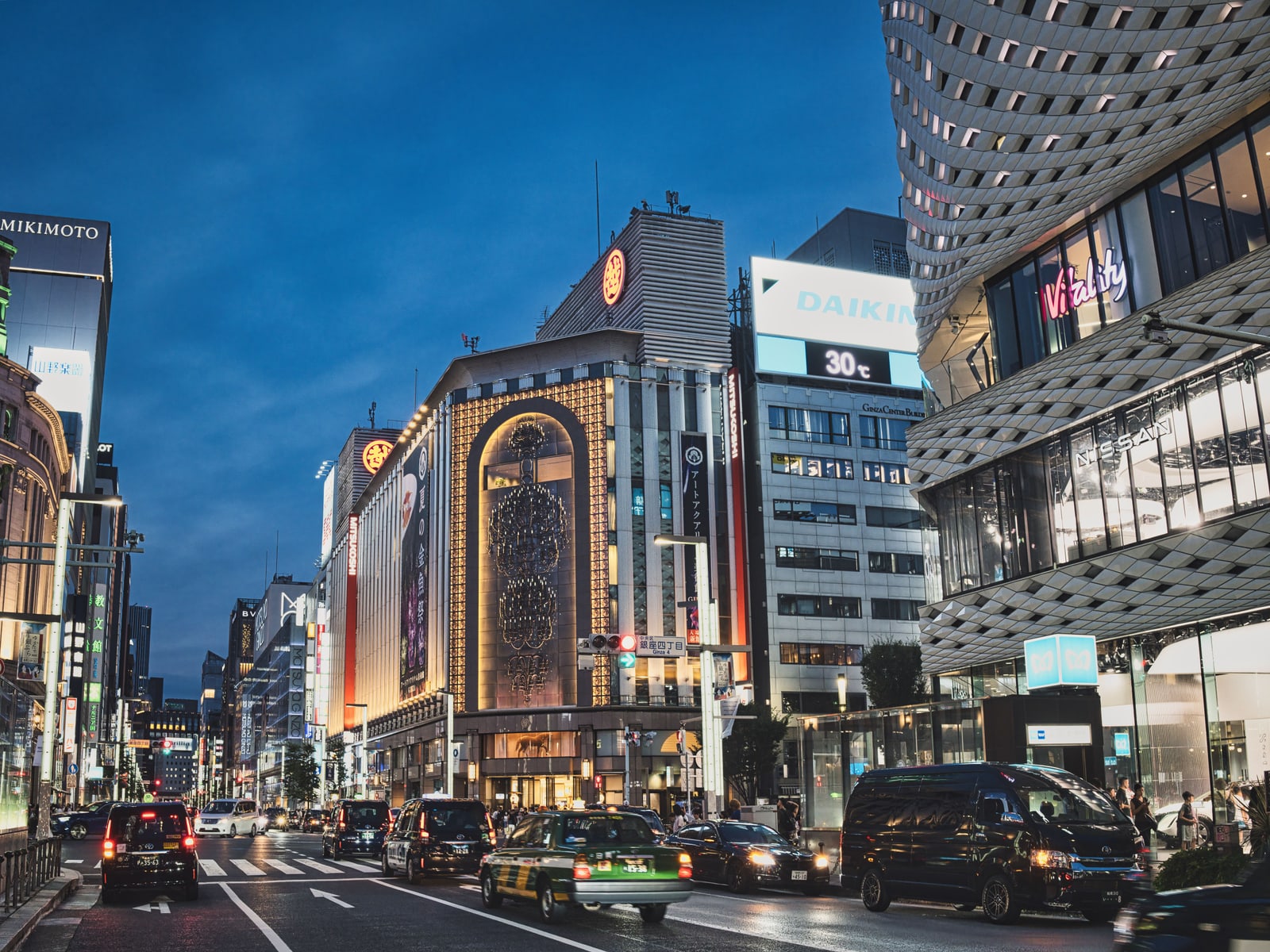 Chuo-dori in Ginza at dusk, a lit department-store facade under a deep blue sky, taxis in the road and a street sign reading Ginza 4