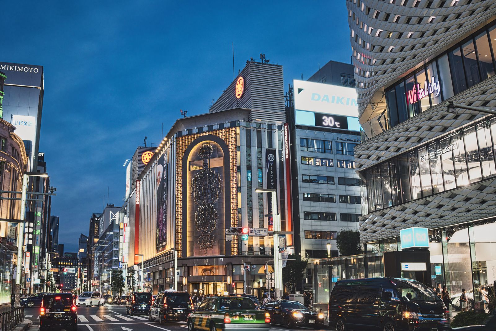 A Ginza crossing at dusk with a Ginza 4-chome street sign, a lit department store with a gold-framed arched window and taxis waiting at the signal