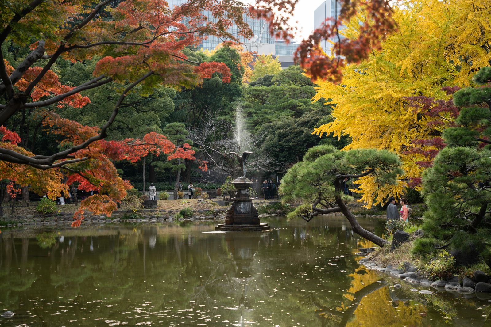 A park pond with a bronze crane fountain among red maples and a yellow ginkgo in autumn, office towers faintly visible behind the trees