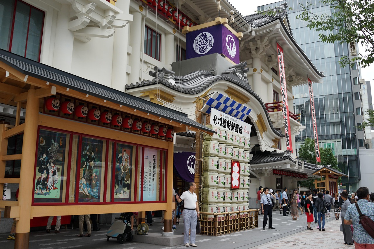 The white curved gable and purple crest curtain of the Kabukiza, a stack of sake barrels and a poster board under red lanterns, passers-by on the pavement
