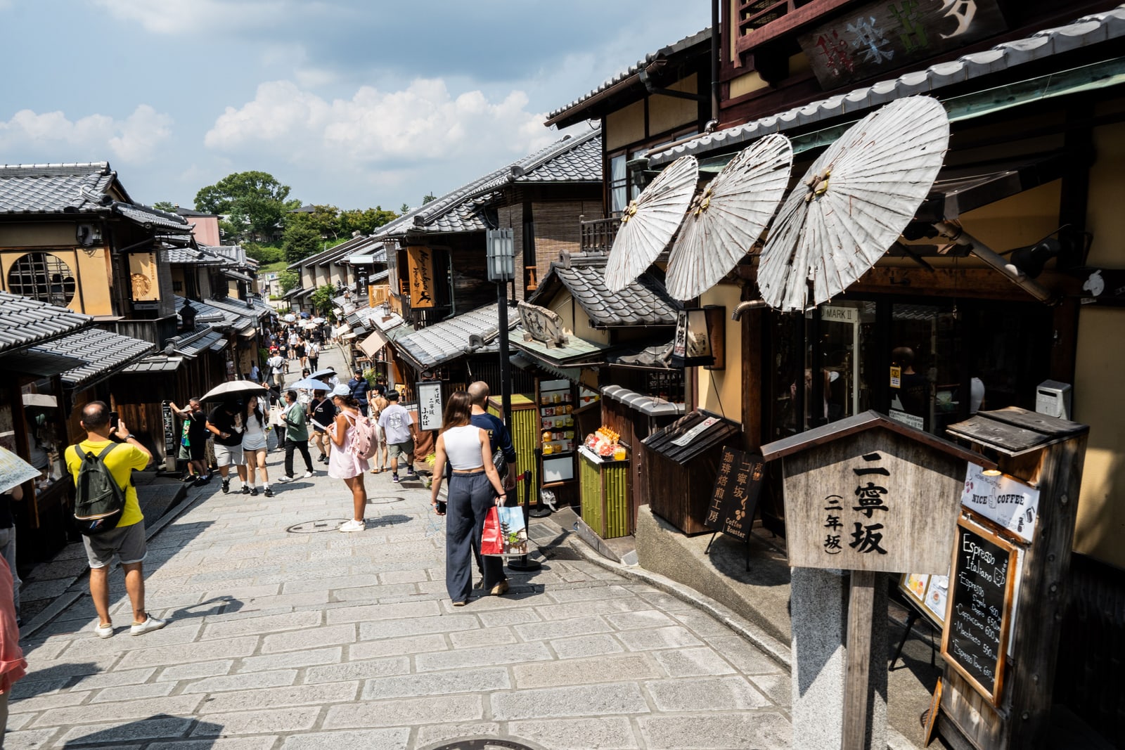A crowd on the stone-paved Ninenzaka slope, wooden shopfronts and three paper parasols at the right