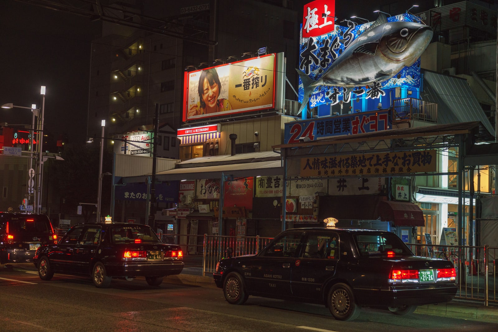 Black taxis on a wet city street at night under a large illuminated fish-shaped restaurant sign.