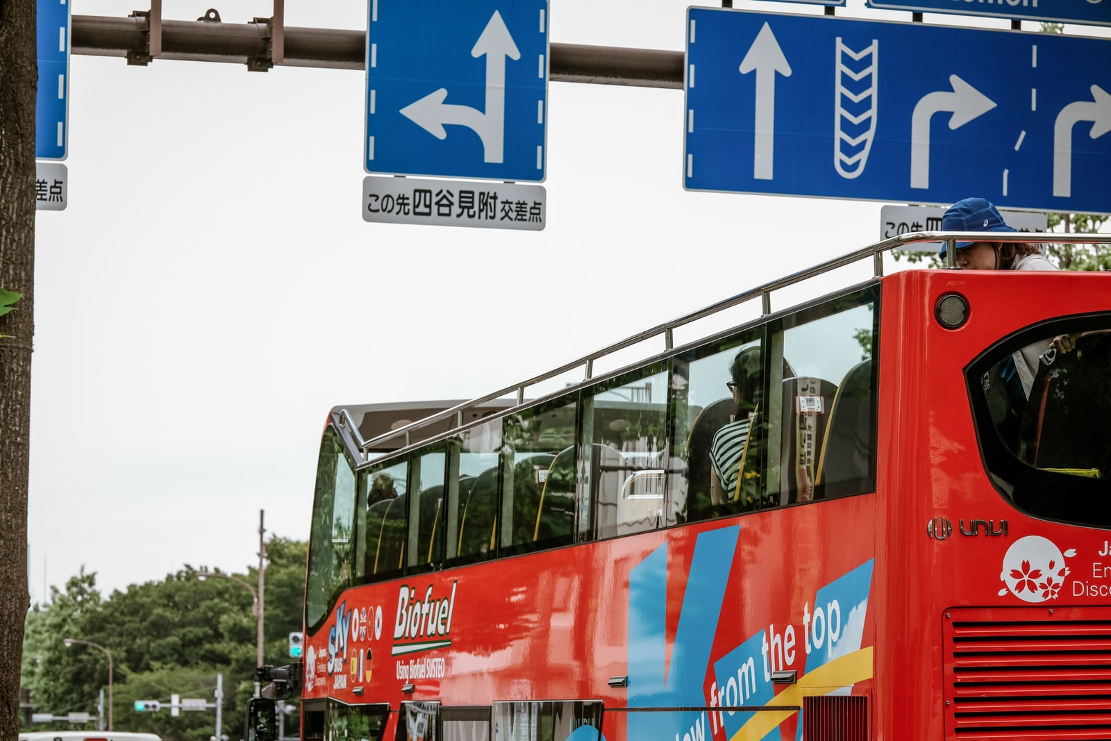 A red open-top double-decker sightseeing bus under blue Japanese road signs, passengers on the upper deck.