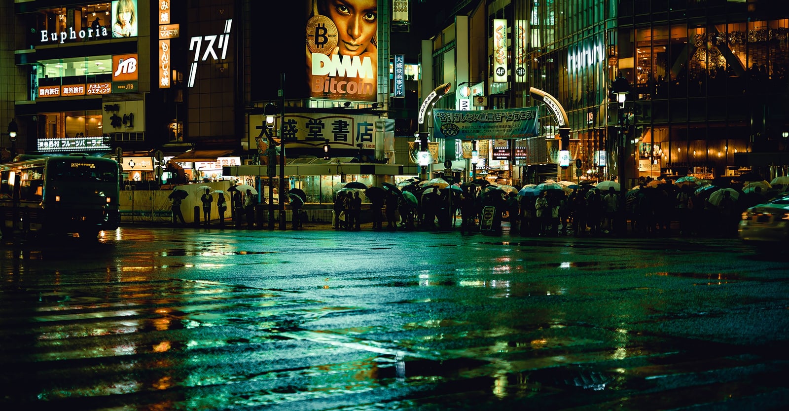 A wide wet intersection at night reflecting green and yellow neon, with people under umbrellas on the far corner.