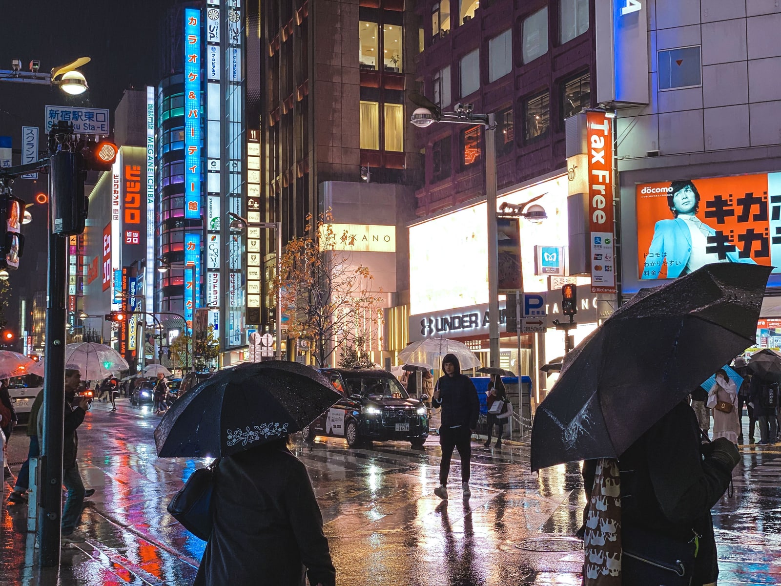 People holding black umbrellas on a rainy shopping street at night, with lit store signs reflected on the pavement.