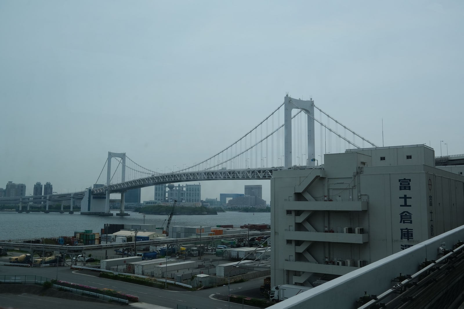 Rainbow Bridge spanning Tokyo Bay under a gray sky, seen past a harbor warehouse roof.