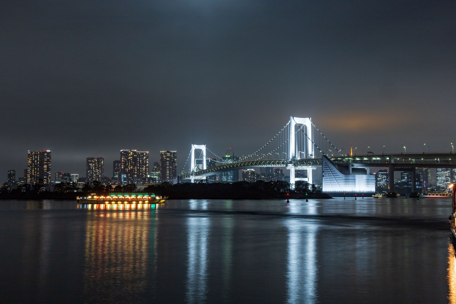 Rainbow Bridge lit white at night across calm water, with the city skyline and a lit boat below.