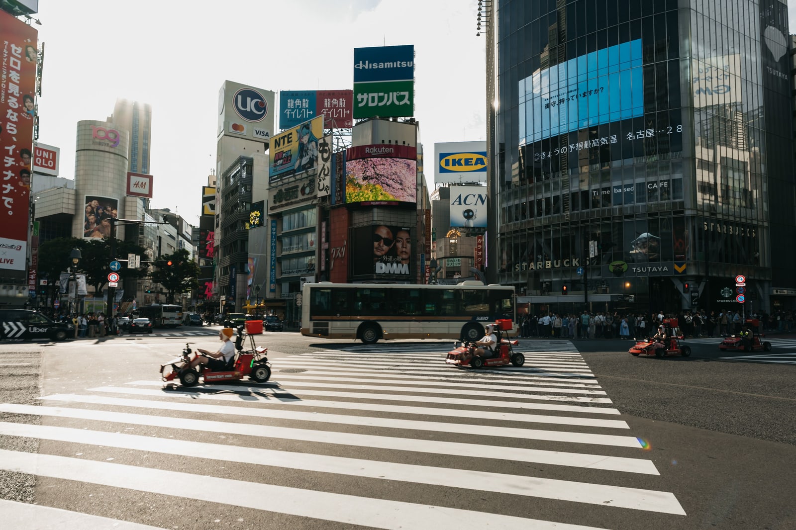 Four small go-karts crossing the zebra stripes of Shibuya Crossing in daylight, with a city bus and billboards behind them.