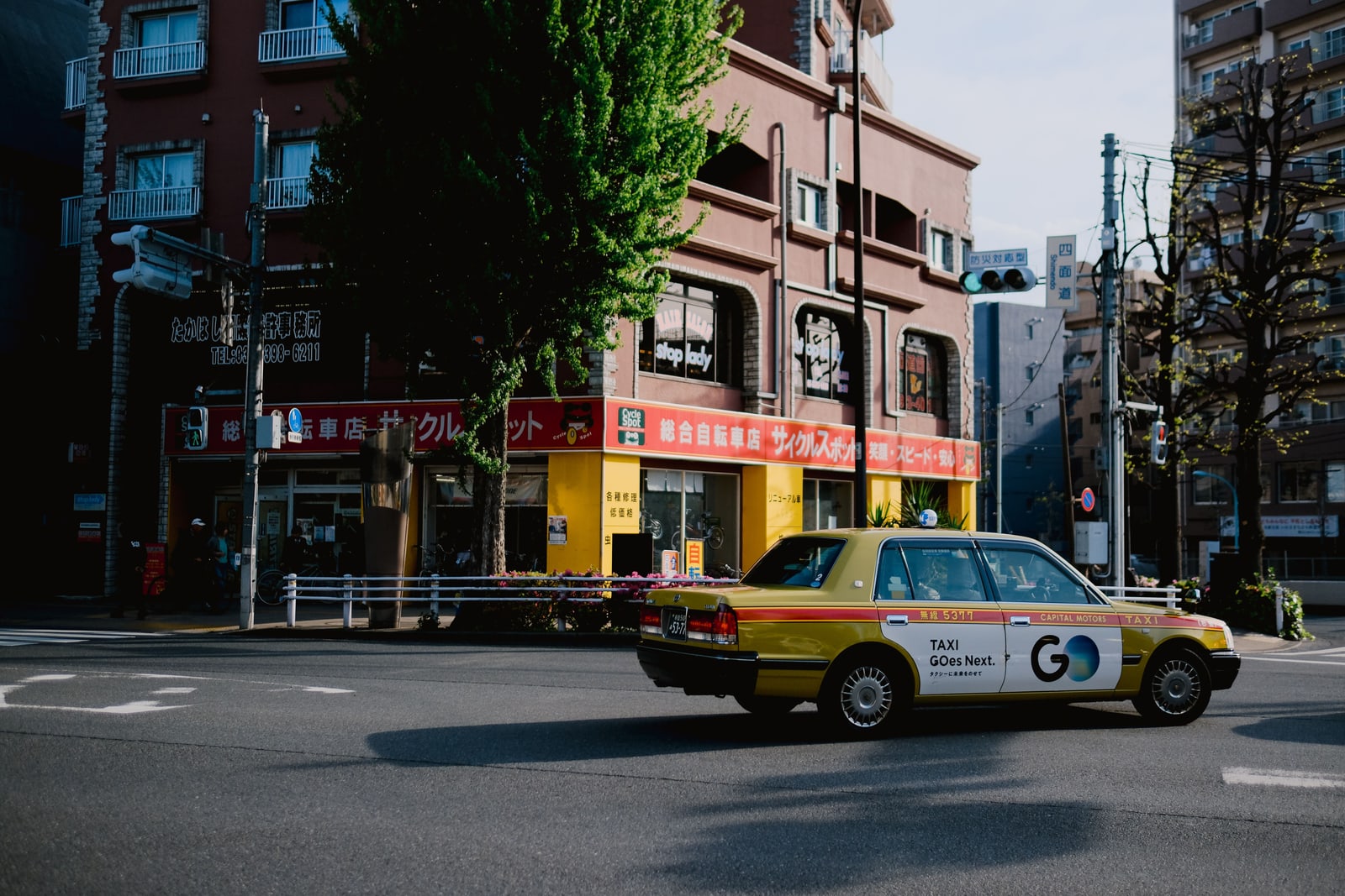 A yellow Tokyo taxi turning at a quiet street corner past shops and a tree.